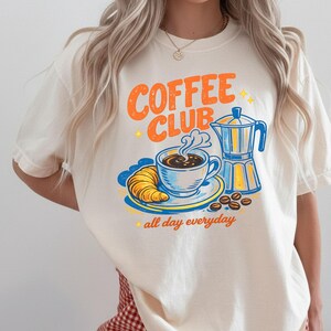 May include: Off-white t-shirt featuring a vintage-style "Coffee Club" design in orange and blue. The graphic includes a coffee cup, croissant, coffee beans, and a coffee pot. The words "all day everyday" are also included.