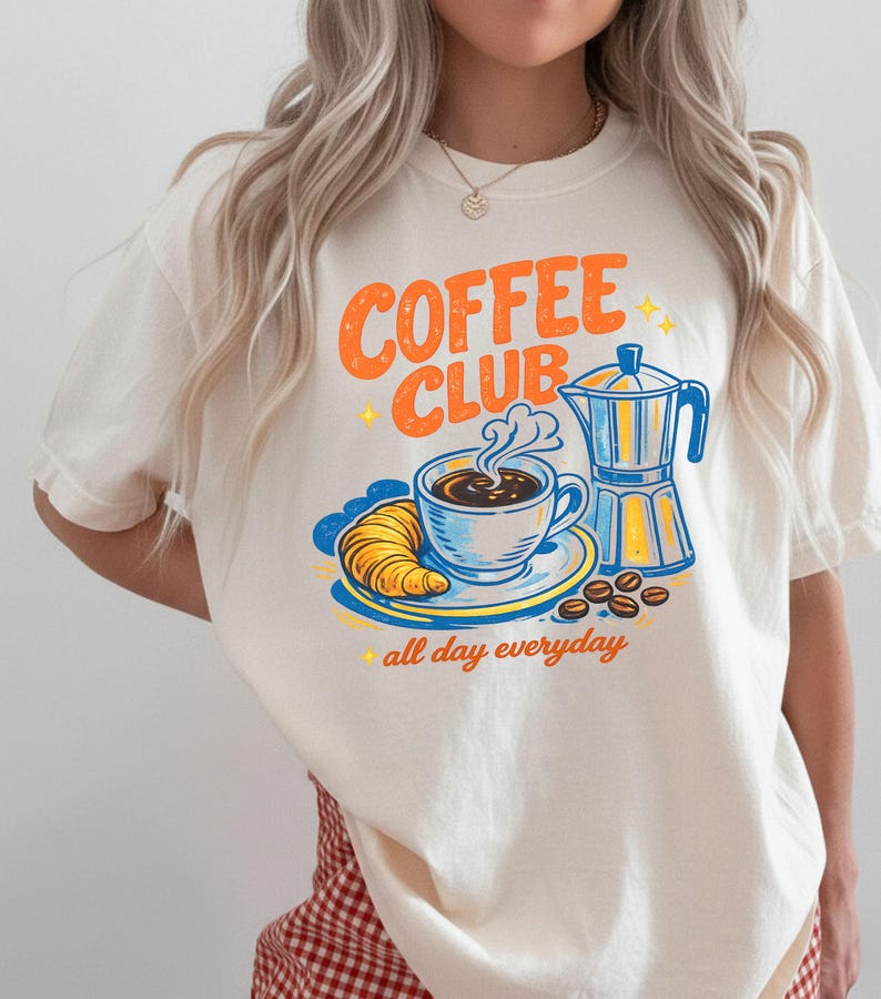 May include: Off-white t-shirt featuring a vintage-style "Coffee Club" design in orange and blue. The graphic includes a coffee cup, croissant, coffee beans, and a coffee pot. The words "all day everyday" are also included.