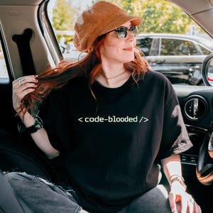 K&ouml;nnte beinhalten: Schwarzes T-Shirt mit dem Text "< code-blooded />" in Wei&szlig;. Die Person im Bild tr&auml;gt einen braunen Cordhut und eine Sonnenbrille. Die Person sitzt in einem Auto.