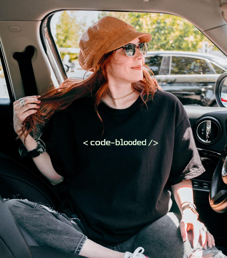 K&ouml;nnte beinhalten: Schwarzes T-Shirt mit dem Text "< code-blooded />" in Wei&szlig;. Die Person im Bild tr&auml;gt einen braunen Cordhut und eine Sonnenbrille. Die Person sitzt in einem Auto.