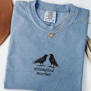 Peut inclure: T-shirt bleu clair avec un motif de deux corbeaux noirs et le texte "attempted murder". Un collier dor&eacute; avec un pendentif rond est pos&eacute; sur le t-shirt. Le t-shirt est pli&eacute; sur une surface blanche.