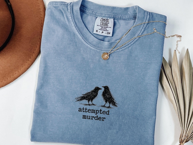 Peut inclure: T-shirt bleu clair avec un motif de deux corbeaux noirs et le texte "attempted murder". Un collier dor&eacute; avec un pendentif rond est pos&eacute; sur le t-shirt. Le t-shirt est pli&eacute; sur une surface blanche.