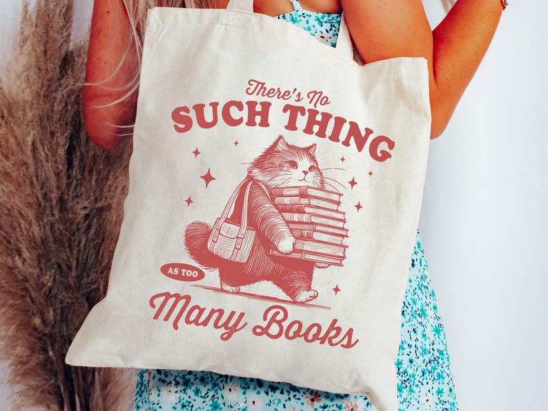 Peut inclure: Sac fourre-tout cr&egrave;me avec une illustration rouge d'un chat portant une pile de livres et un sac. Le texte "There's No Such Thing As Too Many Books" est imprim&eacute; en rouge. Le sac est tenu par une personne.