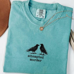 Peut inclure: Un t-shirt turquoise Comfort Colors avec un motif noir de deux corbeaux et le texte "attempted murder". Un collier dor&eacute; avec un pendentif repose sur le t-shirt. Un chapeau marron et des feuilles de palmier s&eacute;ch&eacute;es sont en arri&egrave;re-plan.