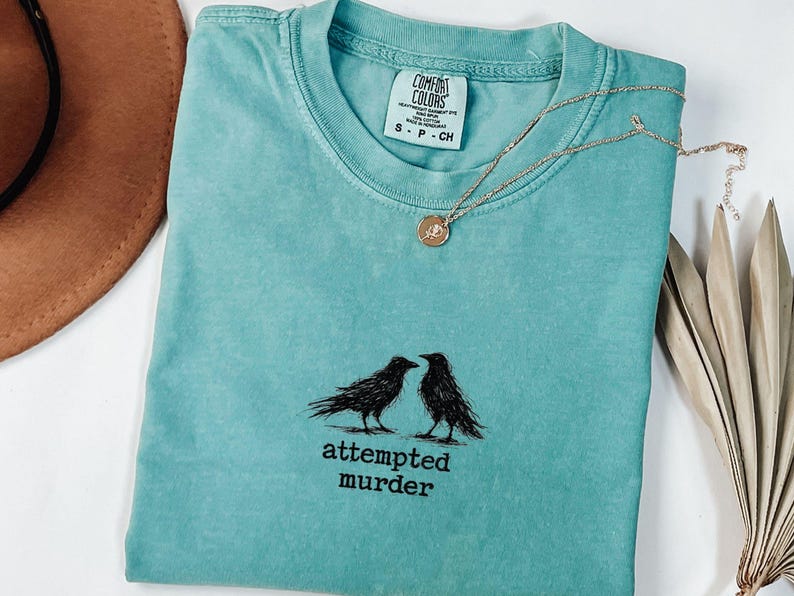 Peut inclure: Un t-shirt turquoise Comfort Colors avec un motif noir de deux corbeaux et le texte "attempted murder". Un collier dor&eacute; avec un pendentif repose sur le t-shirt. Un chapeau marron et des feuilles de palmier s&eacute;ch&eacute;es sont en arri&egrave;re-plan.