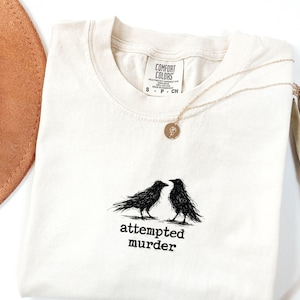Peut inclure: T-shirt cr&egrave;me Comfort Colors avec un motif noir de deux corbeaux se faisant face, avec les mots "attempted murder" en dessous. Un collier en or avec un pendentif est pos&eacute; sur le t-shirt.
