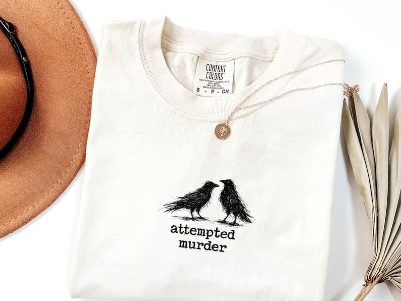 Peut inclure: T-shirt cr&egrave;me Comfort Colors avec un motif noir de deux corbeaux se faisant face, avec les mots "attempted murder" en dessous. Un collier en or avec un pendentif est pos&eacute; sur le t-shirt.