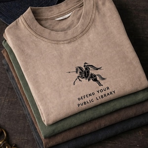 May include: A stack of four t-shirts in various earth tones: tan, olive green, brown, and navy blue. The top shirt features a black graphic of a knight on horseback with the text "DEFEND YOUR PUBLIC LIBRARY" below the image.
