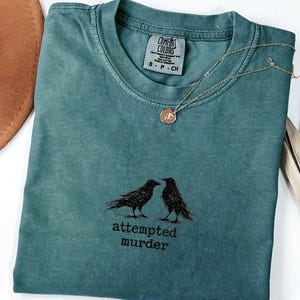 Peut inclure: T-shirt de couleur sarcelle avec un motif de deux corbeaux noirs et le texte "attempted murder". Un collier en or avec un pendentif rond est pos&eacute; sur le t-shirt. Le t-shirt est pli&eacute;, avec un chapeau marron et une plante s&eacute;ch&eacute;e visibles.