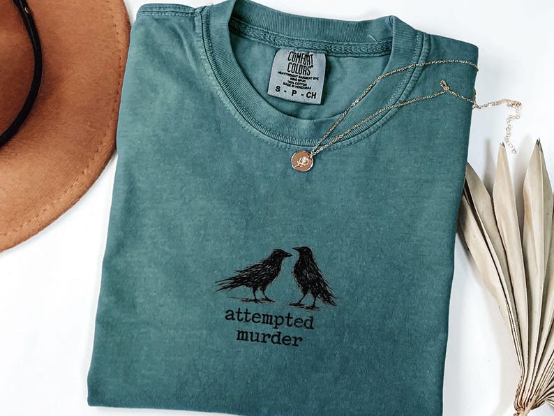 Peut inclure: T-shirt de couleur sarcelle avec un motif de deux corbeaux noirs et le texte "attempted murder". Un collier en or avec un pendentif rond est pos&eacute; sur le t-shirt. Le t-shirt est pli&eacute;, avec un chapeau marron et une plante s&eacute;ch&eacute;e visibles.