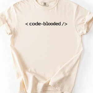 K&ouml;nnte beinhalten: Cremefarbenes T-Shirt mit dem Text "< code-blooded />" in Schwarz. Das Shirt hat einen Rundhalsausschnitt und kurze &Auml;rmel, wobei die &Auml;rmel hochgekrempelt sind. Der Stoff wirkt weich und bequem.