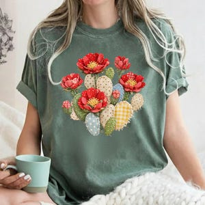 Puede incluir: Camiseta verde con un diseño de cactus con flores rojas de tela y varios detalles de tela. El cactus tiene un diseño de patchwork con lunares y cuadros de vichy. La camiseta es de corte holgado.