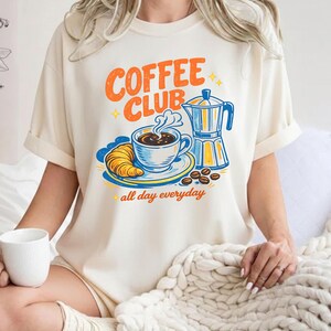 May include: A cream-coloured t-shirt featuring a retro "Coffee Club" design in orange and blue. The graphic includes a coffee cup, croissant, coffee beans, and a coffee maker. The words "all day everyday" are below the graphic.