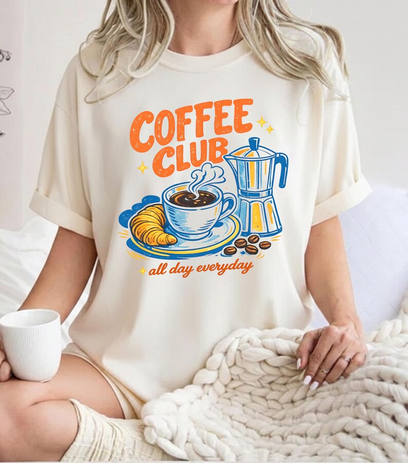 May include: A cream-coloured t-shirt featuring a retro "Coffee Club" design in orange and blue. The graphic includes a coffee cup, croissant, coffee beans, and a coffee maker. The words "all day everyday" are below the graphic.