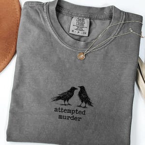 Peut inclure: Un t-shirt gris Comfort Colors avec un motif de deux corbeaux noirs et le texte "attempted murder". Un collier pendentif dor&eacute; et un chapeau marron sont &eacute;galement visibles. Le t-shirt est de taille S-P-CH.
