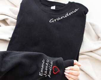May include: Black sweatshirt with the word "Grandma" embroidered in white script. The sleeve features the names "Emma James Amelia" and a red heart. The sweatshirt is folded on a neutral-colored blanket.