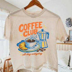 May include: A cream-coloured t-shirt featuring a coffee-themed graphic. The design includes the words "COFFEE CLUB" in orange, a coffee cup, croissant, coffee beans, and a blue and yellow coffee maker. The phrase "all day everyday" is also present.