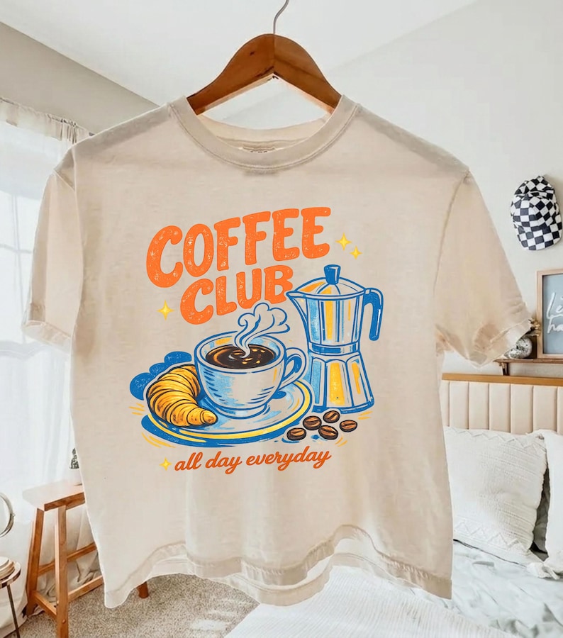 May include: A cream-coloured t-shirt featuring a coffee-themed graphic. The design includes the words "COFFEE CLUB" in orange, a coffee cup, croissant, coffee beans, and a blue and yellow coffee maker. The phrase "all day everyday" is also present.
