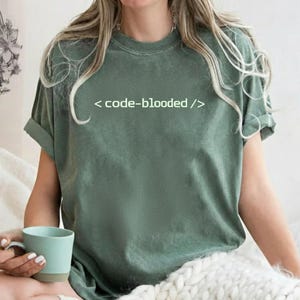 Puede incluir: Camiseta verde salvia con el texto "< code-blooded />" en blanco. La camiseta tiene mangas cortas y un corte relajado. Una persona sostiene una taza azul claro.
