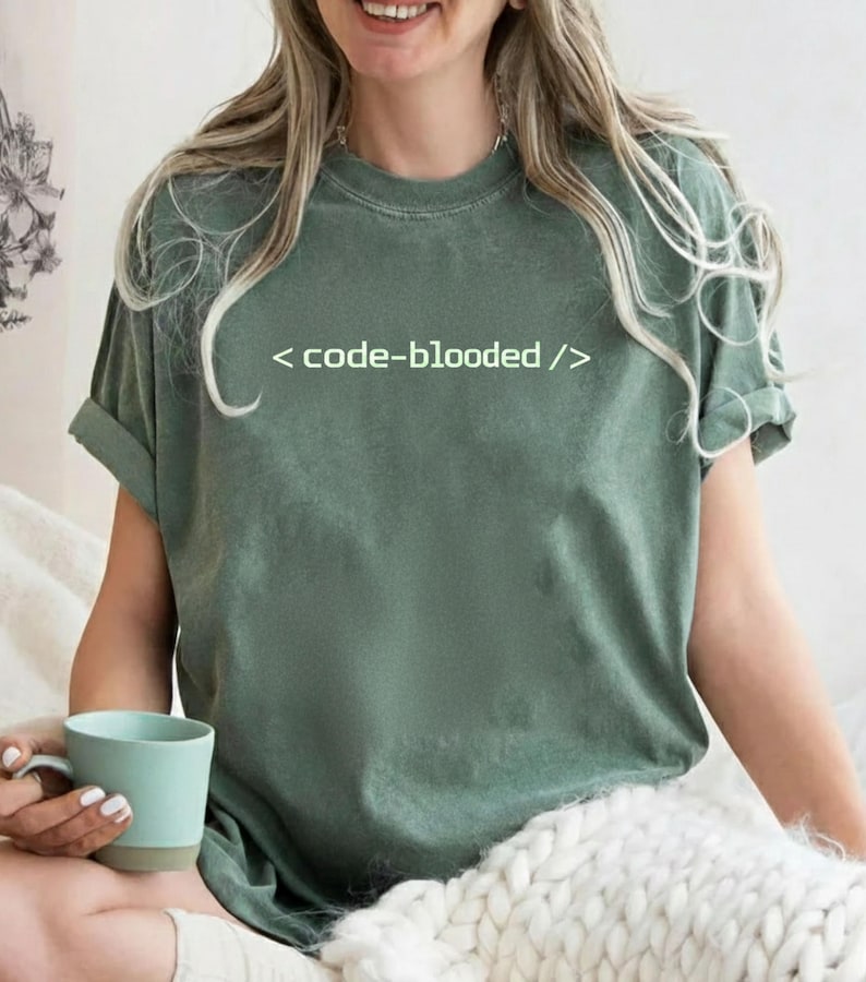 K&ouml;nnte beinhalten: Salbeigr&uuml;nes T-Shirt mit dem Text "< code-blooded />" in Wei&szlig;. Das Shirt hat kurze &Auml;rmel und eine lockere Passform. Eine Person h&auml;lt einen hellblauen Becher.