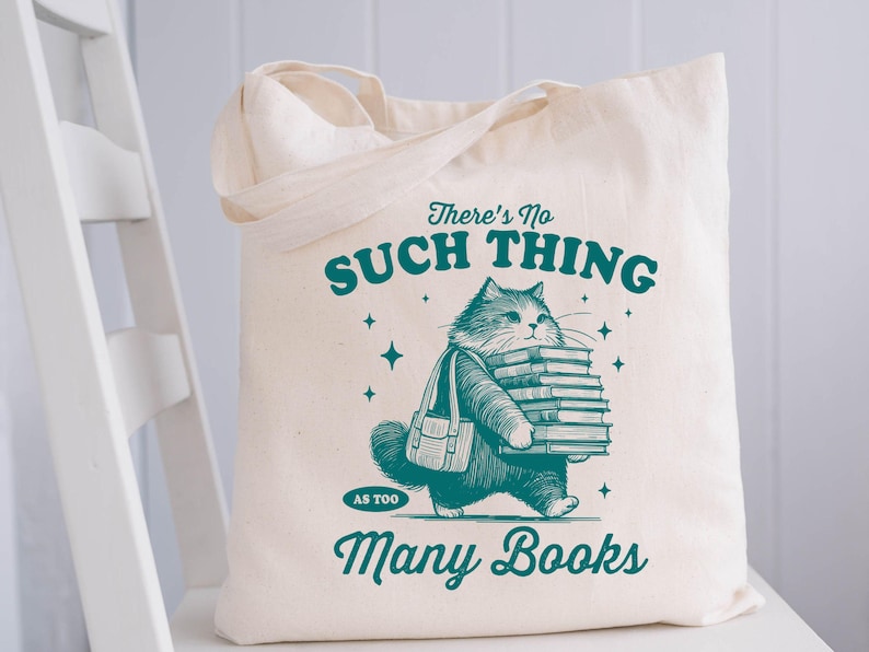 Peut inclure: Sac fourre-tout cr&egrave;me avec une illustration turquoise d'un chat portant des livres. Le texte sur le sac dit "There's No Such Thing As Too Many Books". Le sac est rectangulaire avec deux anses.