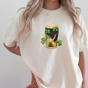 Puede incluir: Camiseta color crema con un gráfico de un vaso de cerveza decorado con patrones de patchwork y tréboles. El diseño del vaso incorpora telas verdes, azul marino y amarillas. La camiseta tiene mangas cortas y cuello redondo.