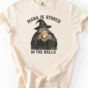 Könnte beinhalten: Cremefarbenes T-Shirt mit einer Grafik eines Zauberers, der eine leuchtende Kugel hält. Der Text "MANA IS STORED IN THE BALLS" ist über und unter der Zauberer-Illustration aufgedruckt. Das Shirt hat kurze Ärmel und einen Rundhalsausschnitt.