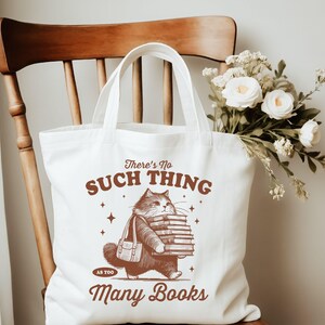 Peut inclure: Un sac fourre-tout blanc avec une illustration marron d'un chat portant une pile de livres. Le texte sur le sac se lit &laquo; There's No Such Thing As Too Many Books &raquo;. Le sac est pos&eacute; sur une chaise en bois, avec un bouquet de fleurs blanches.
