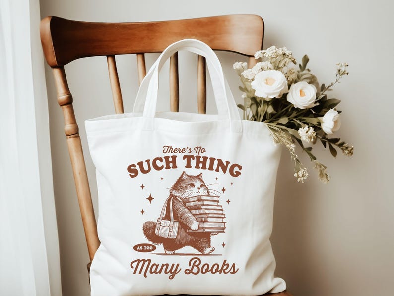 Peut inclure: Un sac fourre-tout blanc avec une illustration marron d'un chat portant une pile de livres. Le texte sur le sac se lit &laquo; There's No Such Thing As Too Many Books &raquo;. Le sac est pos&eacute; sur une chaise en bois, avec un bouquet de fleurs blanches.