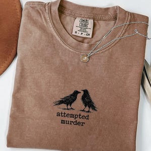 Peut inclure: T-shirt marron Comfort Colors &agrave; col rond, avec un motif noir de deux corbeaux et le texte "attempted murder". Un collier argent&eacute; avec un pendentif repose sur le t-shirt. Un chapeau marron est visible en arri&egrave;re-plan.