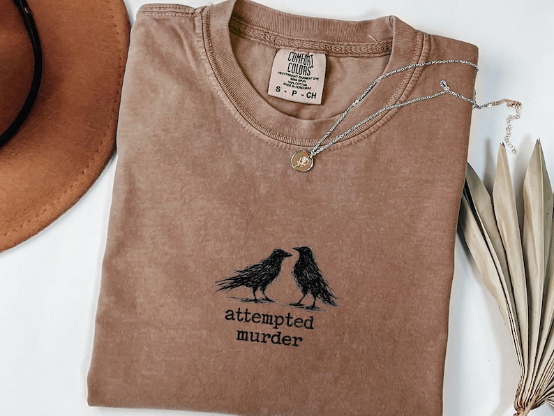 Peut inclure: T-shirt marron Comfort Colors &agrave; col rond, avec un motif noir de deux corbeaux et le texte "attempted murder". Un collier argent&eacute; avec un pendentif repose sur le t-shirt. Un chapeau marron est visible en arri&egrave;re-plan.