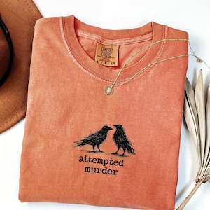Peut inclure: T-shirt corail &agrave; col rond, avec un motif noir de deux corbeaux et le texte "attempted murder". Un collier dor&eacute; avec un pendentif et un chapeau marron sont &eacute;galement visibles.