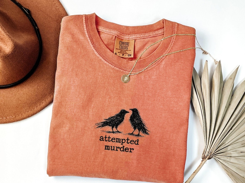Peut inclure: T-shirt corail &agrave; col rond, avec un motif noir de deux corbeaux et le texte "attempted murder". Un collier dor&eacute; avec un pendentif et un chapeau marron sont &eacute;galement visibles.