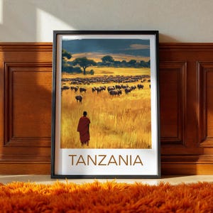 Puede incluir: Impresión artística enmarcada que presenta una escena de paisaje vibrante. Una persona con una prenda roja se encuentra en hierba alta y dorada, frente a una manada de animales. La palabra "TANZANIA" está impresa en marrón en la parte inferior.