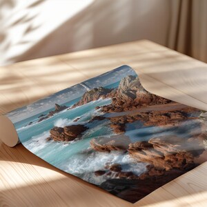May include: A rolled print depicting a coastal scene with turquoise water and reddish-brown rocks. The print is on a wooden surface, with sunlight and shadows enhancing the image. The artwork captures the essence of the ocean and natural landscapes.