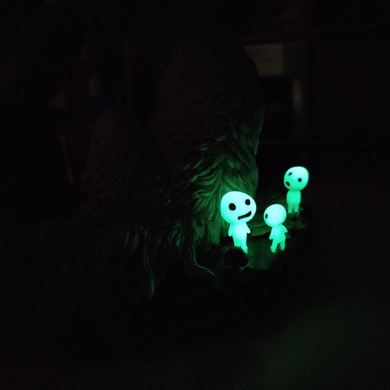 May include: Glow-in-the-dark figurines of small, humanoid forest spirits with simple facial features. The figures are a bright green color and appear to be standing near a dark, textured object, possibly a tree or rock formation. The scene is set against a black background.