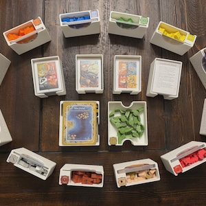May include: A collection of white plastic board game organizers containing various colored wooden tokens, card decks, and game components. The set includes multiple rectangular trays with cutouts, holding orange, blue, green, yellow, purple, black, grey, red, and tan pieces, along with illustrated game cards.