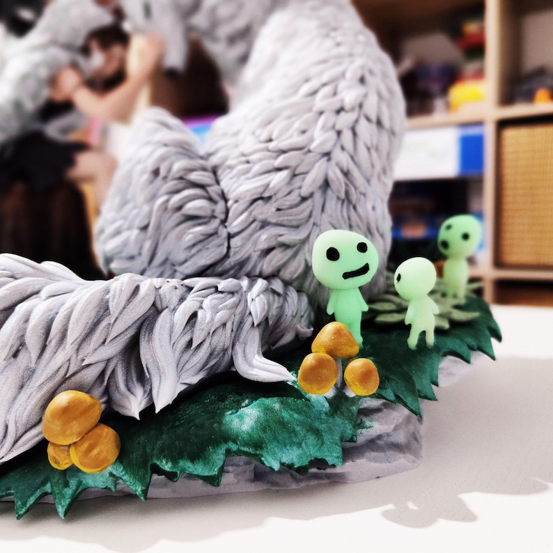 May include: A detailed sculpture featuring a large, textured, gray and white creature with a fluffy appearance. Small green figures with black facial features stand near yellow mushrooms and green foliage. The sculpture is on a white surface.