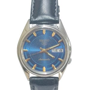 Pode incluir: Um relógio de pulso automático Seiko vintage com mostrador azul e marcadores de hora dourados. O relógio tem uma caixa prateada e uma pulseira de couro preta. O dia é exibido como "TUE 8".