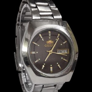 K&ouml;nnte beinhalten: Silberfarbene Orient-Uhr mit dunkelbraunem Zifferblatt. Die Uhr hat goldfarbene Zeiger und Markierungen sowie eine Tagesdatumsanzeige. Das Armband besteht aus silberfarbenen Metallgliedern. Eine klassische Uhr.