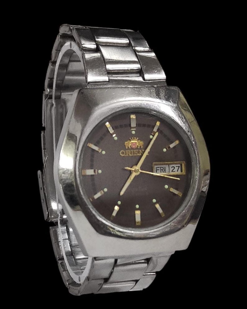 K&ouml;nnte beinhalten: Silberfarbene Orient-Uhr mit dunkelbraunem Zifferblatt. Die Uhr hat goldfarbene Zeiger und Markierungen sowie eine Tagesdatumsanzeige. Das Armband besteht aus silberfarbenen Metallgliedern. Eine klassische Uhr.