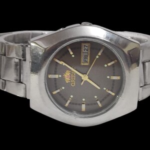 K&ouml;nnte beinhalten: Eine silberfarbene Orient-Uhr mit dunkelbraunem Zifferblatt und goldenen Zeigern und Markierungen. Die Uhr hat eine Tages- und Datumsanzeige und ein silbernes Metallarmband. Das Zifferblatt ist von einer silbernen L&uuml;nette umgeben.