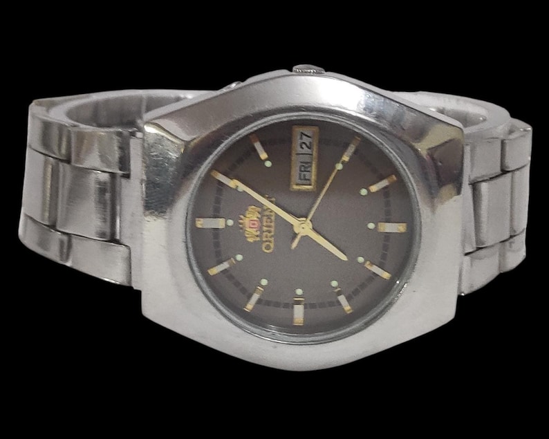 K&ouml;nnte beinhalten: Eine silberfarbene Orient-Uhr mit dunkelbraunem Zifferblatt und goldenen Zeigern und Markierungen. Die Uhr hat eine Tages- und Datumsanzeige und ein silbernes Metallarmband. Das Zifferblatt ist von einer silbernen L&uuml;nette umgeben.