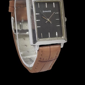 Puede incluir: Un reloj Sonata rectangular con esfera negra y marco plateado. El reloj tiene una correa de cuero marr&oacute;n. La esfera del reloj tiene marcadores y manecillas blancas. La palabra "SONATA" est&aacute; impresa en la esfera.