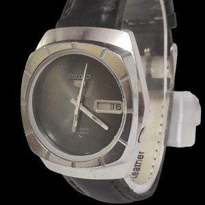 May include: A vintage Seiko 5 automatic wristwatch with a dark gray face and a silver-tone case. The watch features a day and date display and is set on a black leather strap. The watch face displays the number 16.