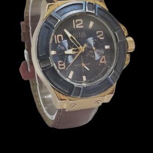 Peut inclure: Une montre Guess dor&eacute;e avec un cadran bleu fonc&eacute; et noir et un bracelet en cuir marron. La montre a un cadran rond avec des aiguilles et des index dor&eacute;s. Le bracelet pr&eacute;sente un motif textur&eacute; en forme de crocodile.