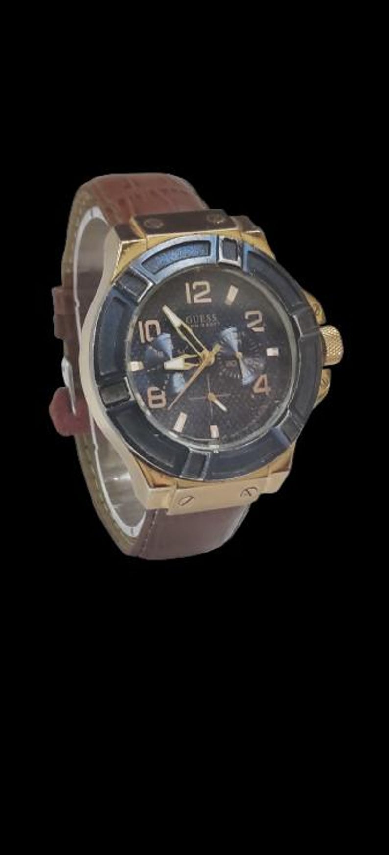 Peut inclure: Une montre Guess dor&eacute;e avec un cadran bleu fonc&eacute; et noir et un bracelet en cuir marron. La montre a un cadran rond avec des aiguilles et des index dor&eacute;s. Le bracelet pr&eacute;sente un motif textur&eacute; en forme de crocodile.
