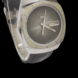May include: A vintage Seiko 5 automatic watch with a black leather band. The watch has a silver-colored case and a dark gray face with a date display. The watch hands and markers are silver.