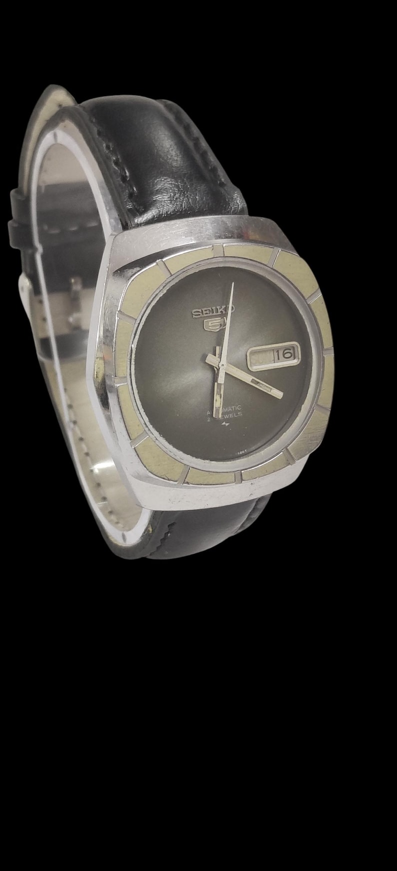 May include: A vintage Seiko 5 automatic watch with a black leather band. The watch has a silver-colored case and a dark gray face with a date display. The watch hands and markers are silver.