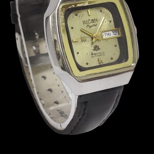 May include: A vintage Ricoh Crystal automatic wristwatch with a gold-toned square face and black leather strap. The watch face displays the day and date, with the text "THU 12". The watch has a silver-toned case.